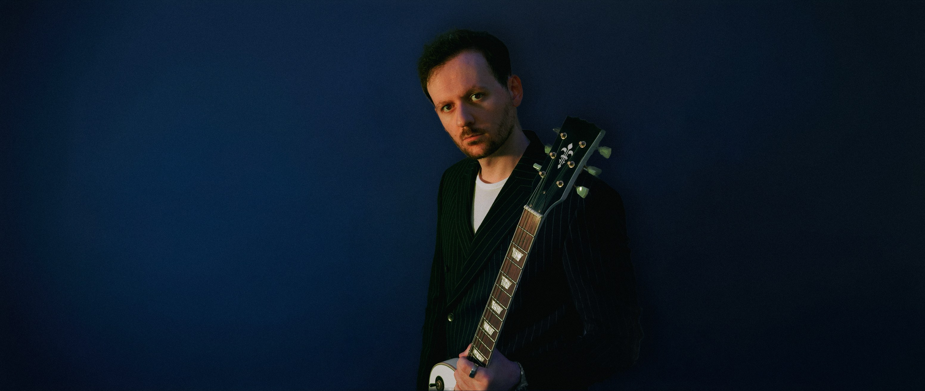 Fraid in a pinstripe suit against a dramatic navy background, holding a guitar by the headstock